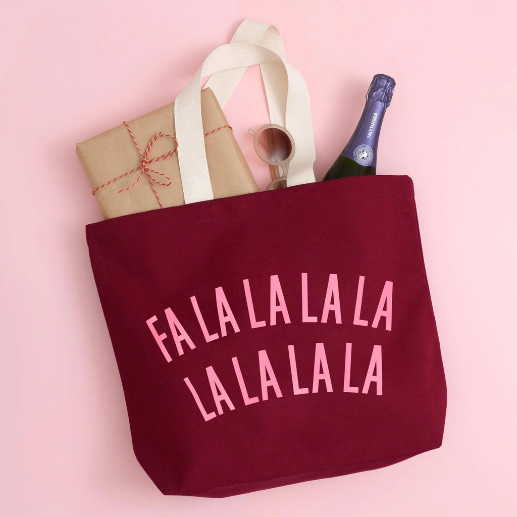 A burgundy canvas tote bag with cream handles and pink text reading 'FALALALALALA' across the front, shown on a pink background with a wrapped gift box and a champagne bottle tucked inside.