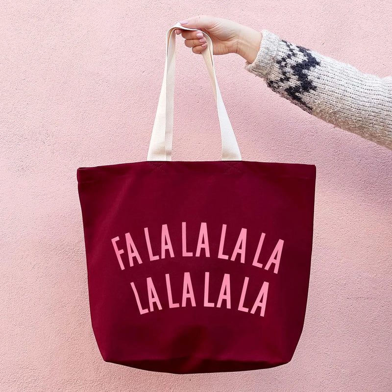 A burgundy canvas tote bag with white handles and pink text reading 'FALALALALA LA LA LA LA LA' on the front, held by a hand wearing a knit sweater against a pink wall.