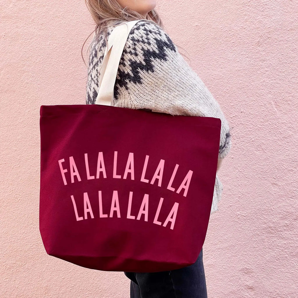 A person wearing a cream and black patterned knit sweater carries a large burgundy canvas tote bag with the phrase 'FALALALALALA' printed in light pink letters across the front, set against a textured pink wall.