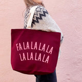 A person wearing a cream and black patterned knit sweater carries a large burgundy canvas tote bag with the phrase 'FALALALALALA' printed in light pink letters across the front, set against a textured pink wall.