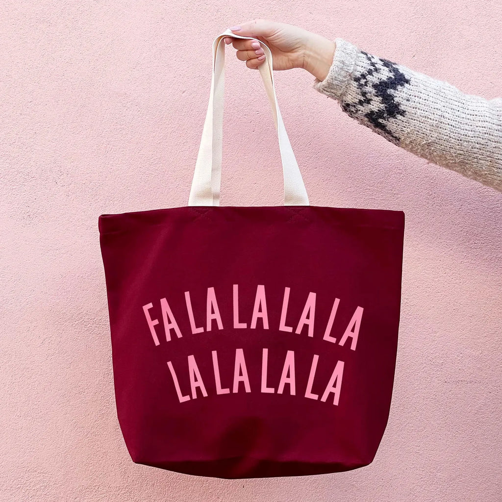 A burgundy canvas tote bag with white handles and pink text reading 'FALALALALA LA LA LA LA LA' on the front, held by a hand wearing a knit sweater against a pink wall.