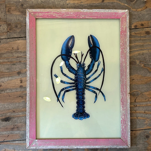 A framed lobster art piece featuring a blue lobster illustration on a cream background with a distressed pink frame, placed on a rustic wooden floor.