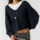 Free People See Me V-Neck pullover. A woman modeling a dark navy blue knit sweater with a wide V-neck and voluminous sleeves, layered over a white top and paired with a silver necklace and a black sequined skirt. 