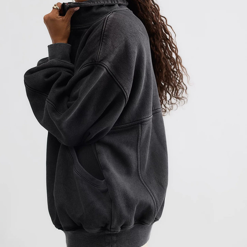 A woman wearing a dark washed black oversized pullover hoodie, shown from a side-back angle against a plain light gray studio background.