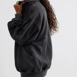 A woman wearing a dark washed black oversized pullover hoodie, shown from a side-back angle against a plain light gray studio background.