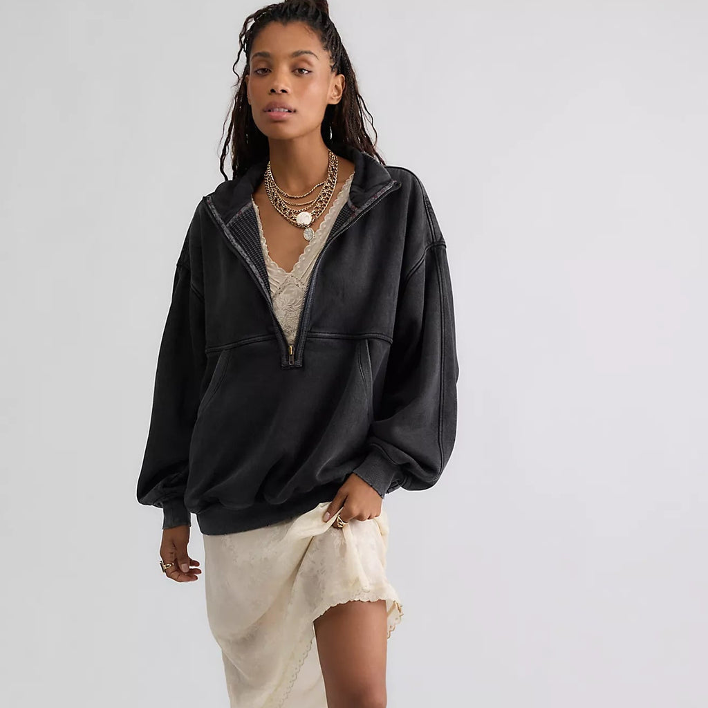 A model stands against a light gray studio background wearing an oversized black zip-up hoodie, a cream asymmetrical mini skirt, layered gold necklaces, and tall black boots.