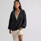 A model stands against a light gray studio background wearing an oversized black zip-up hoodie, a cream asymmetrical mini skirt, layered gold necklaces, and tall black boots.