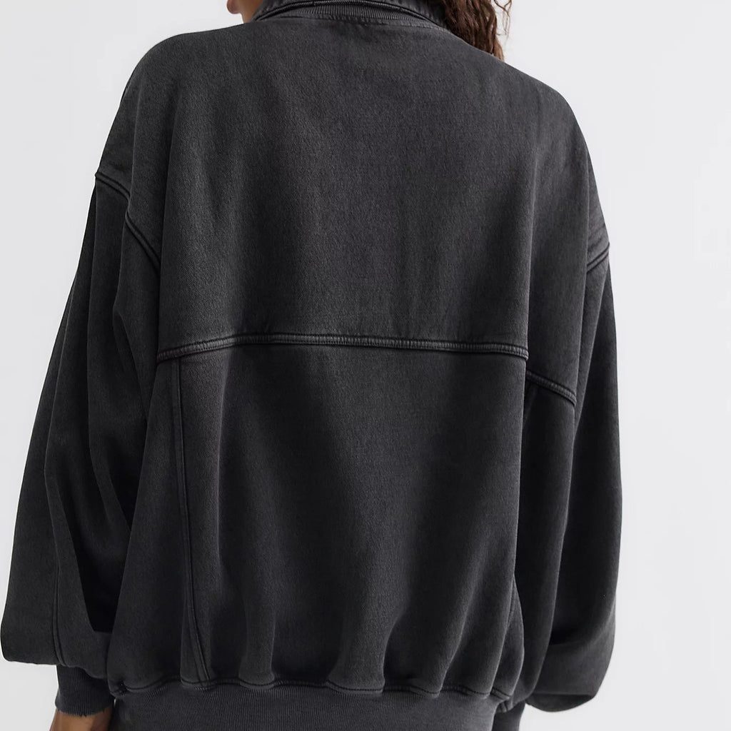 A woman shown from the back wearing a dark washed black oversized bomber-style sweatshirt, paired with light beige pants, against a plain light gray studio background.