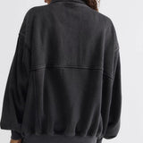 A woman shown from the back wearing a dark washed black oversized bomber-style sweatshirt, paired with light beige pants, against a plain light gray studio background.