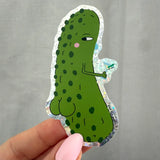 A hand with pink nail polish holds a die-cut sticker of a green cartoon pickle character with a holographic silver border, set against a gray fabric background.