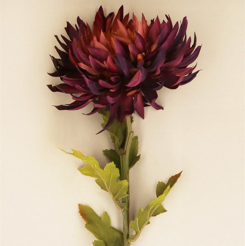 A single artificial chrysanthemum flower with deep burgundy-purple petals and green leaves on a stem, photographed against a plain off-white background.