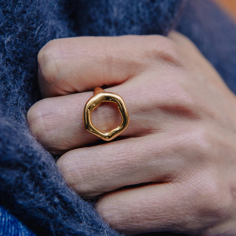 A close-up of a hand wearing a textured, organic-shaped gold ring, nestled against a dark blue fuzzy knit fabric.
