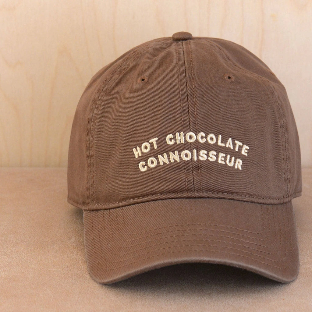 A brown baseball cap with 'HOT CHOCOLATE CONNOISSEUR' embroidered in cream on the front, set against a light wood background.