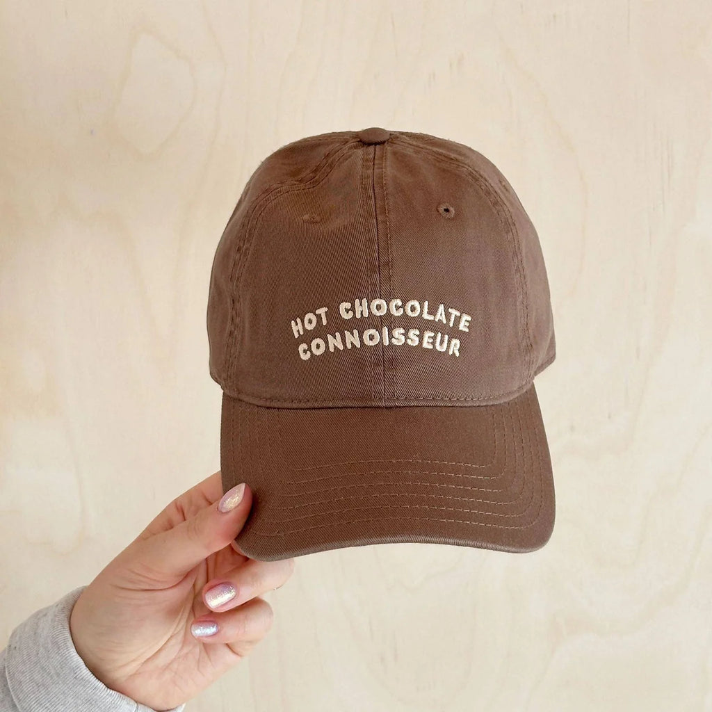 Brown cap with 'Hot Chocolate Connoisseur' text held by a hand against a beige background