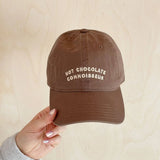 Brown cap with 'Hot Chocolate Connoisseur' text held by a hand against a beige background
