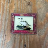 A small framed vintage-style illustration of a white swan standing in water, mounted on a rustic wooden surface. The frame is ornate and pinkish-red with gold detailing.