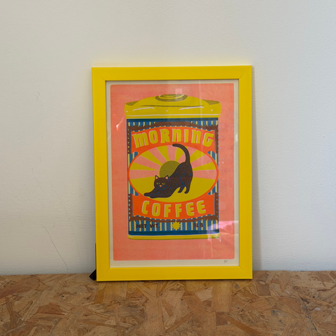 Framed artwork with a black cat design on a pink background, placed on a wooden floor against a white wall.