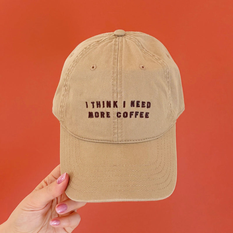 Beige cap with 'I think I need more coffee' text held against a red background