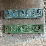 Two rectangular decorative metal door knockers with embossed floral and geometric patterns, showing a green verdigris patina, mounted on a concrete surface outdoors.