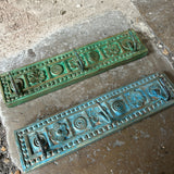 Two ornate rectangular decorative wall plaques with embossed floral and scroll motifs, one in green and one in distressed turquoise-blue, displayed on a concrete surface.