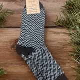 A pair of men's socks with a herringbone pattern, primarily in black and grey, displayed on a wooden surface with Christmas decorations in the background.
