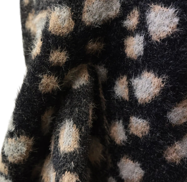 Close-up of a fuzzy knit fabric with a black base and irregular beige and light gray spots, shown with soft folds.