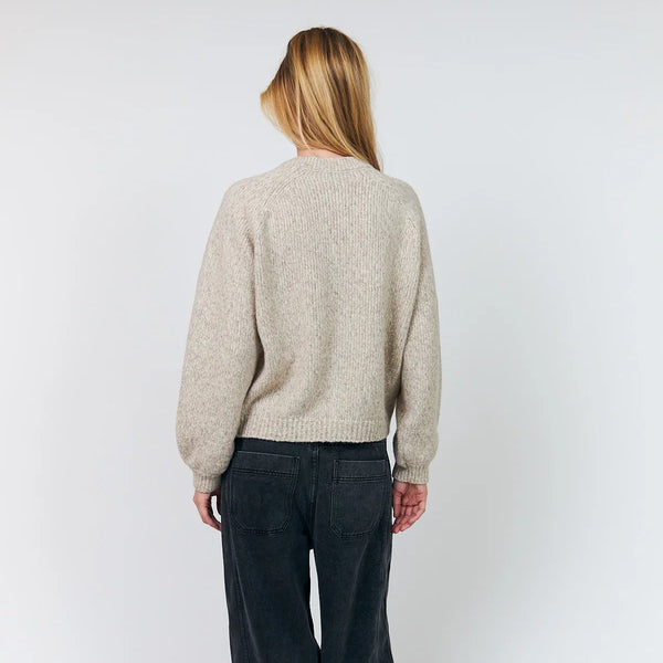 A woman viewed from behind wearing a light beige knit sweater and dark charcoal wide-leg corduroy pants, standing against a plain light gray studio background.