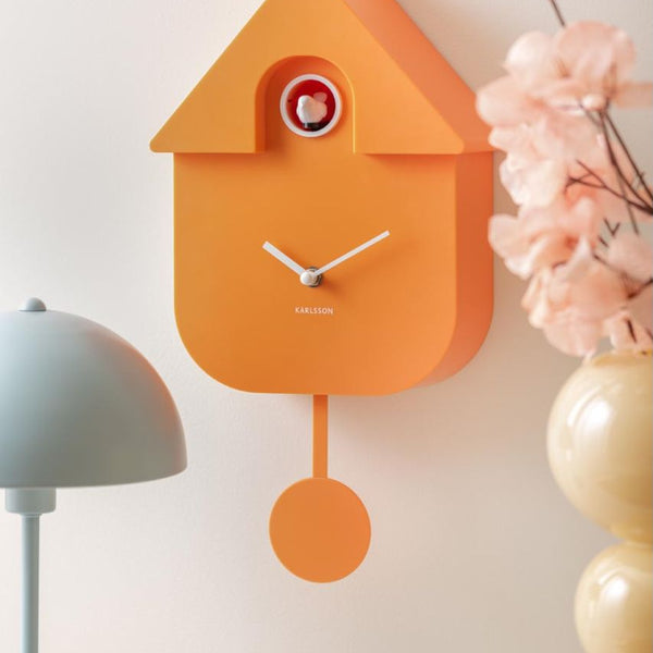 An orange cuckoo clock shaped like a birdhouse with white hands and a small red-and-white bird window, mounted on a light wall beside a gray lamp and pink flowers.
