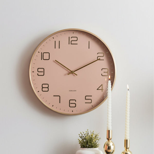 Wall clock with a pink face and gold hands on a white wall above a shelf with decorative items.