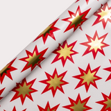 Lagom Design White wrapping paper with a repeating pattern of red and gold starburst shapes, shown partially rolled on a light gray background.