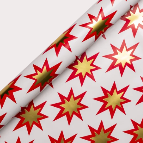 Lagom Design White wrapping paper with a repeating pattern of red and gold starburst shapes, shown partially rolled on a light gray background.