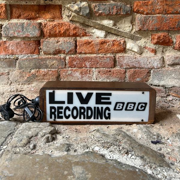 A rectangular wooden box with a white front panel reading 'LIVE RECORDING BBC' in black letters, placed on a stone surface against a brick wall. A coiled black cable lies to the left.