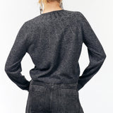 A woman viewed from the back wearing a dark charcoal gray knit sweater with subtle dotted and dashed geometric patterns across the upper back and shoulders, paired with dark gray jeans.