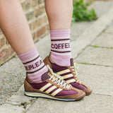 A person wearing pink and maroon crew socks with text and brown low-top sneakers with cream stripes, standing on a paved outdoor surface.