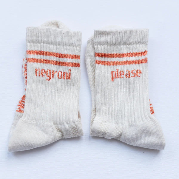 A pair of off-white crew socks with orange stripes and text reading 'negrói' and 'please' on the front, displayed flat against a light background.