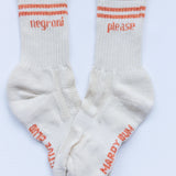 A pair of off-white crew socks with orange stripes and text on the cuffs and soles. One sock has 'negroni' and the other has 'please' printed on the upper cuff, with 'MARDY SUM' printed on the sole.