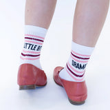 White crew socks with pink and burgundy stripes and the words 'LITTLE BIT OF DRAMA' printed on them, worn with reddish-brown flat shoes against a white background.