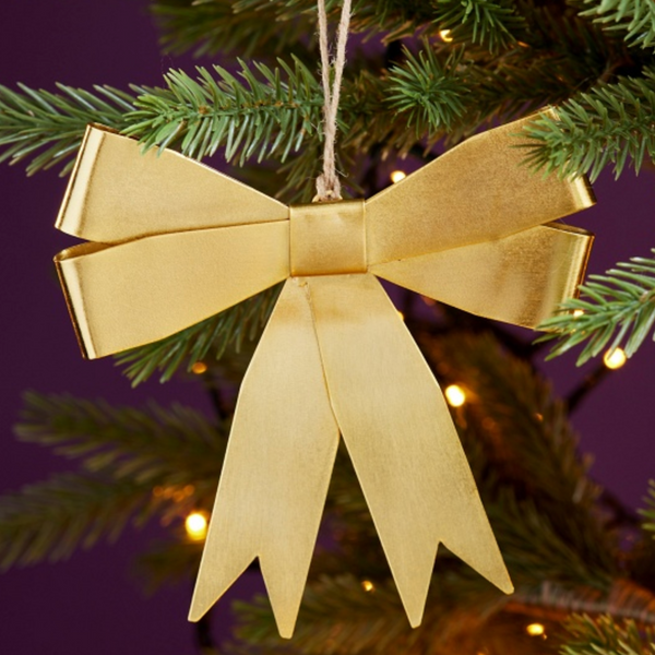 A shiny gold bow Christmas ornament hanging from a green Christmas tree with warm string lights against a dark purple background.