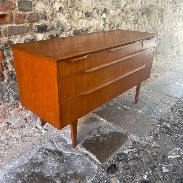 Mid-century modern wooden chest of drawers with three drawers and tapered legs, positioned on a stone floor against a rustic stone wall.