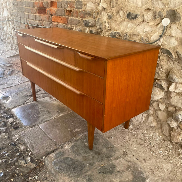 Mid-century modern wooden chest of drawers with three drawers and tapered legs, positioned outdoors against a stone wall and cobblestone ground.