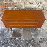 Mid-century modern wooden sideboard with a warm reddish-brown finish, featuring two drawers with curved front panels and tapered legs, set against a rustic stone wall and stone floor.