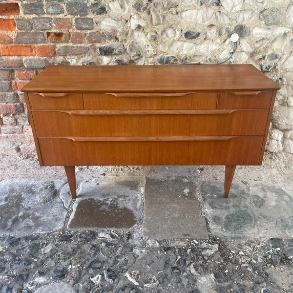 Mid-century modern wooden sideboard with three drawers and tapered legs, photographed outdoors against a stone and brick wall.