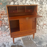 Mid-century modern wooden writing desk cabinet with open shelving, a pull-out desktop, two lower drawers, and a lockable door, shown against a rustic stone and brick wall.