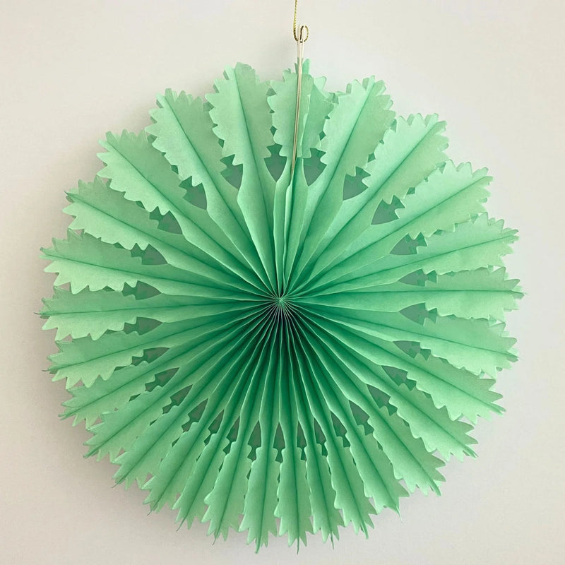A mint green paper fan decoration hanging against a plain off-white wall, with a thin gold hanging string at the top.
