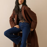 A woman models a stylish outfit consisting of a brown double-breasted coat, dark blue flared jeans, a brown plaid shirt, and a white turtleneck, sitting on a wooden stool against a solid beige background.