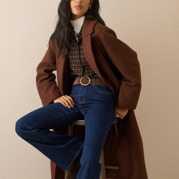 A woman models a stylish outfit consisting of a brown double-breasted coat, dark blue flared jeans, a brown plaid shirt, and a white turtleneck, sitting on a wooden stool against a solid beige background.