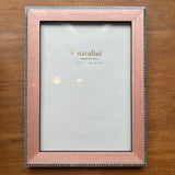 A rectangular picture frame with a light pink wood finish and a silver beaded border, shown empty on a wooden tabletop. The insert card is visible inside the frame, showing the brand name 'natalini'.