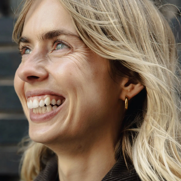 Close-up of a woman with blonde hair and a gold earring, smiling outdoors.