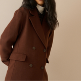 A woman wearing a double-breasted brown wool coat over a cream turtleneck sweater, photographed against a solid beige studio background.