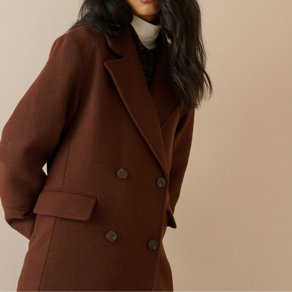 A woman wearing a double-breasted brown wool coat over a cream turtleneck sweater, photographed against a solid beige studio background.